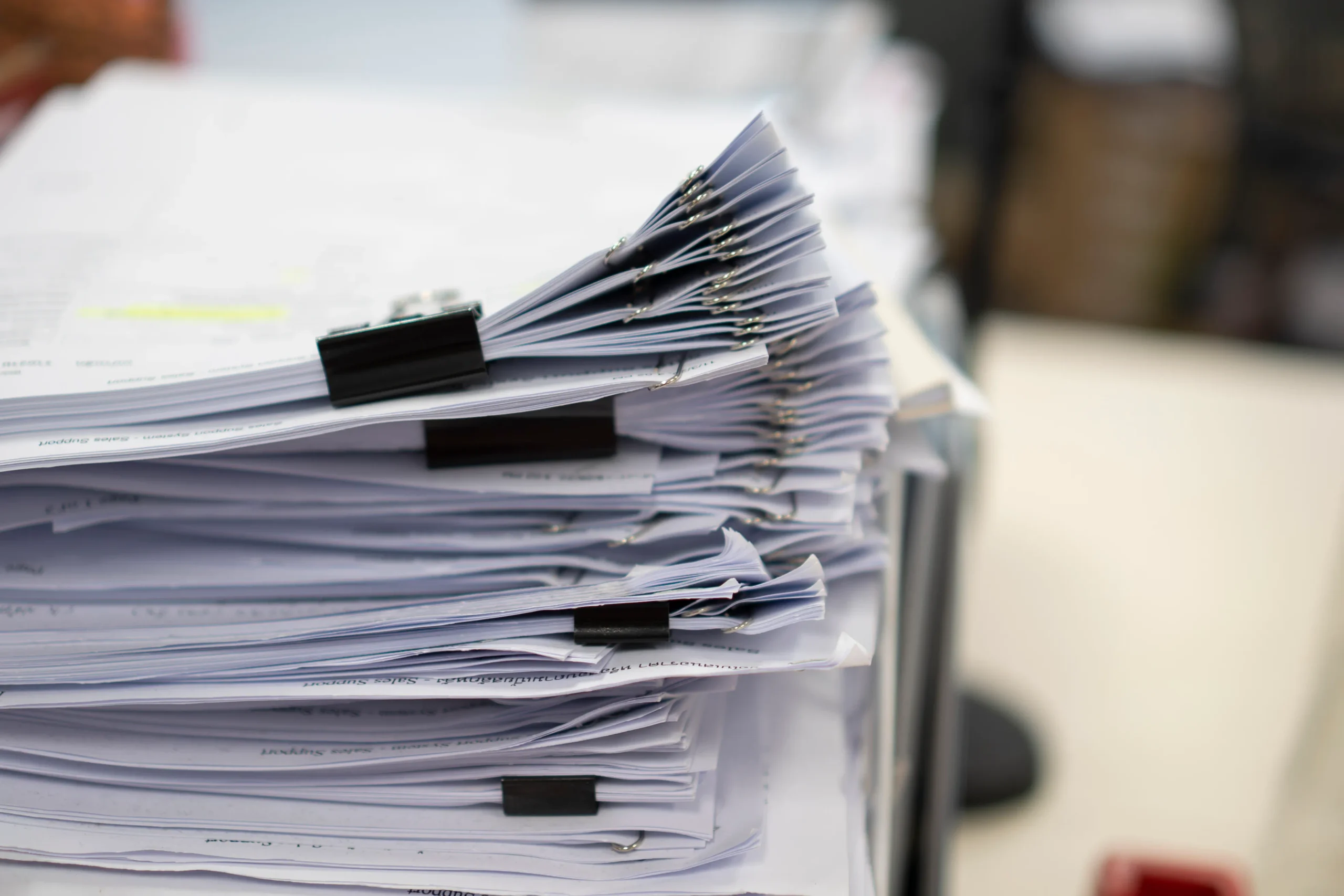 Thick stacks of paper documents held together with binder clips piling up on an office desk, waiting for manual data entry instead of document processing automation